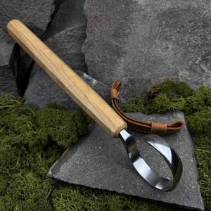 May include: A wooden handled leather carving tool with a curved blade. The tool is resting on a rock with moss growing around it.