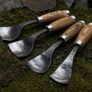 May include: Four wood-handled carving tools with metal blades. The blades are curved and have a spoon-like shape. The tools are arranged on a bed of green moss.