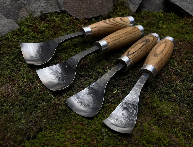 May include: Four wood-handled carving tools with metal blades. The blades are curved and have a spoon-like shape. The tools are arranged on a bed of green moss.