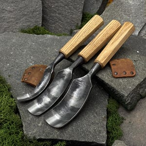 May include: Three hand-forged metal gouges with curved, dark grey steel blades and light brown wooden handles. Each tool features a distinct width and a curved edge, accompanied by small, square brown leather protective covers with metal rivets.