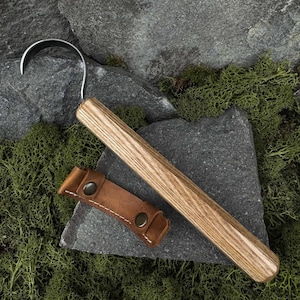 May include: A handcrafted, curved blade tool with a wooden handle and a leather sheath. The blade is made of polished metal. The handle is made of light brown wood. The leather sheath is brown with two metal snaps.