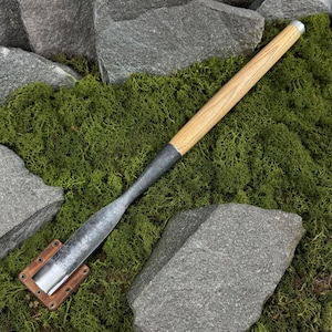 Puede incluir: Un cincel artesanal con un mango de madera clara, un eje de metal oscuro y una base de metal color cobre. El cincel se encuentra sobre un fondo de musgo verde y rocas grises, resaltando su estética rústica.