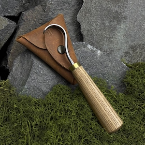 May include: A handcrafted leather sheath holds a hook-shaped tool with a wooden handle and brass accents. The tool's hook is silver-colored. The sheath is brown and secured with a dark button. The handle has a wood grain pattern.
