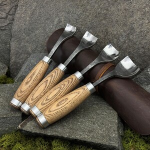 May include: A set of four metal skiving tools with light brown wood handles and silver-colored metal ferrules. Each tool features a curved, wide, polished steel blade designed for leatherworking, arranged in a row with uniform construction.
