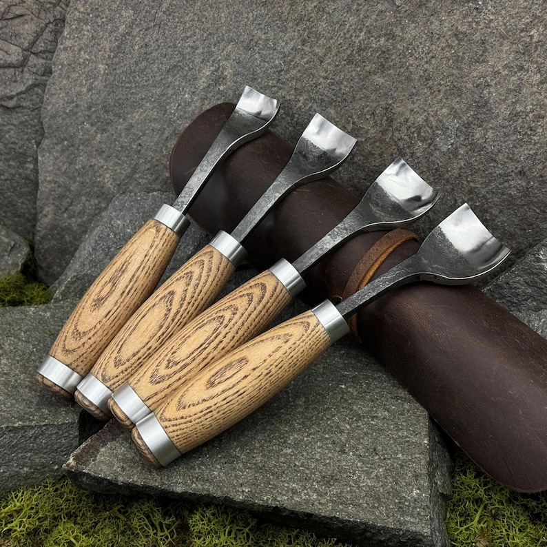 May include: A set of four metal skiving tools with light brown wood handles and silver-colored metal ferrules. Each tool features a curved, wide, polished steel blade designed for leatherworking, arranged in a row with uniform construction.