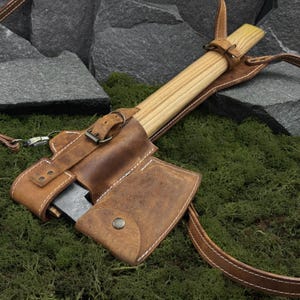 May include: A small axe with a wooden handle and a leather sheath. The sheath is brown with white stitching and a buckle closure. The axe is resting on a bed of green moss.