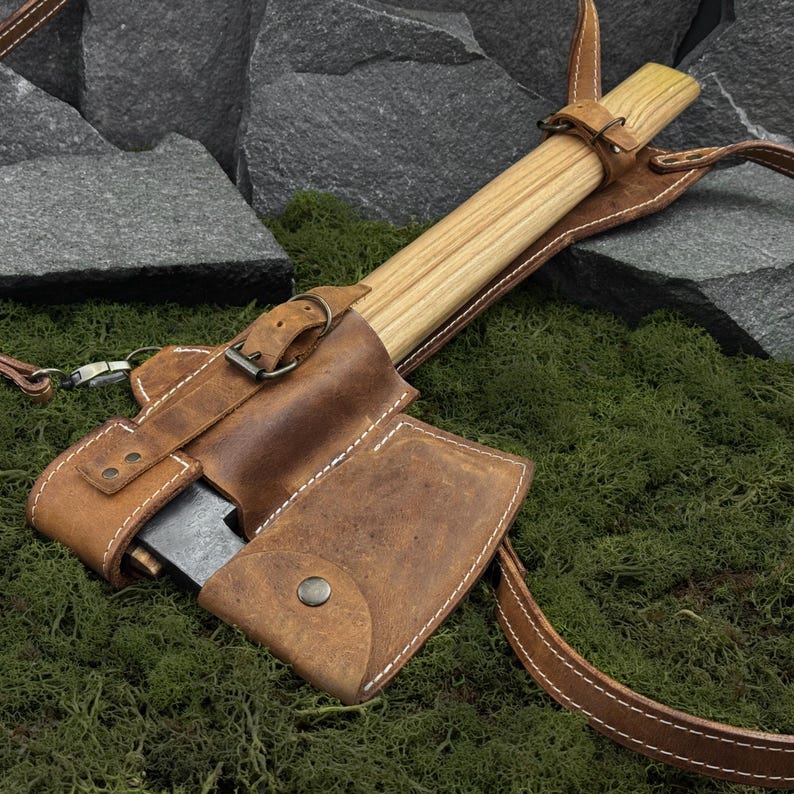 May include: A small axe with a wooden handle and a leather sheath. The sheath is brown with white stitching and a buckle closure. The axe is resting on a bed of green moss.