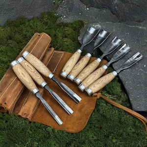 May include: A set of wood carving chisels with wooden handles and metal blades. The chisels are arranged on a brown leather tool roll, set against a backdrop of green moss and dark gray rocks. The tools are designed for woodworking.