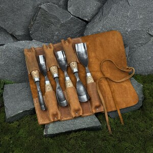 May include: A set of five wood carving gouges with polished metal blades and light-colored wooden handles, organized in a tan leather tool roll. The roll features individual stitched pockets for each tool and a long leather tie for secure closure.