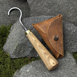 May include: A handcrafted, curved blade tool with a wooden handle and brass ferrule, accompanied by a brown leather pouch. The tool's blade is silver, and the handle has a natural wood grain finish. The leather pouch has a snap closure.