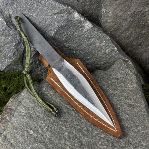 May include: A handmade throwing knife with a sharp, silver blade and a dark, textured handle. It is nestled in a brown leather sheath with white stitching. A green cord is attached to the handle. The knife is displayed on a gray rock.