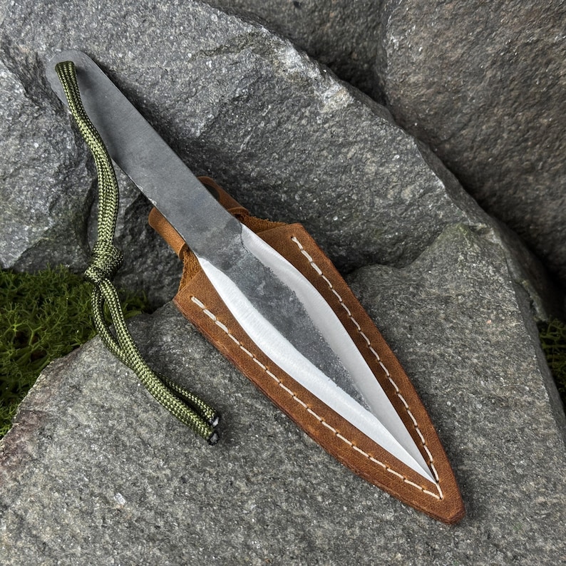 May include: A handmade throwing knife with a sharp, silver blade and a dark, textured handle. It is nestled in a brown leather sheath with white stitching. A green cord is attached to the handle. The knife is displayed on a gray rock.
