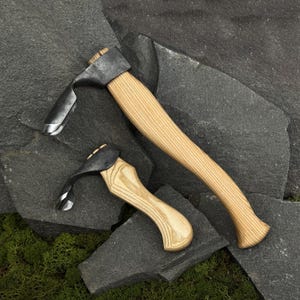 Op de afbeelding: Twee handgereedschappen met gebogen, lichtgekleurde houten handvatten en donkere, gesmede metalen koppen. Elk gereedschap heeft een opvallend, scherp, gebogen blad dat uit de metalen kop steekt, ontworpen voor snij- of houtbewerkingsklussen.