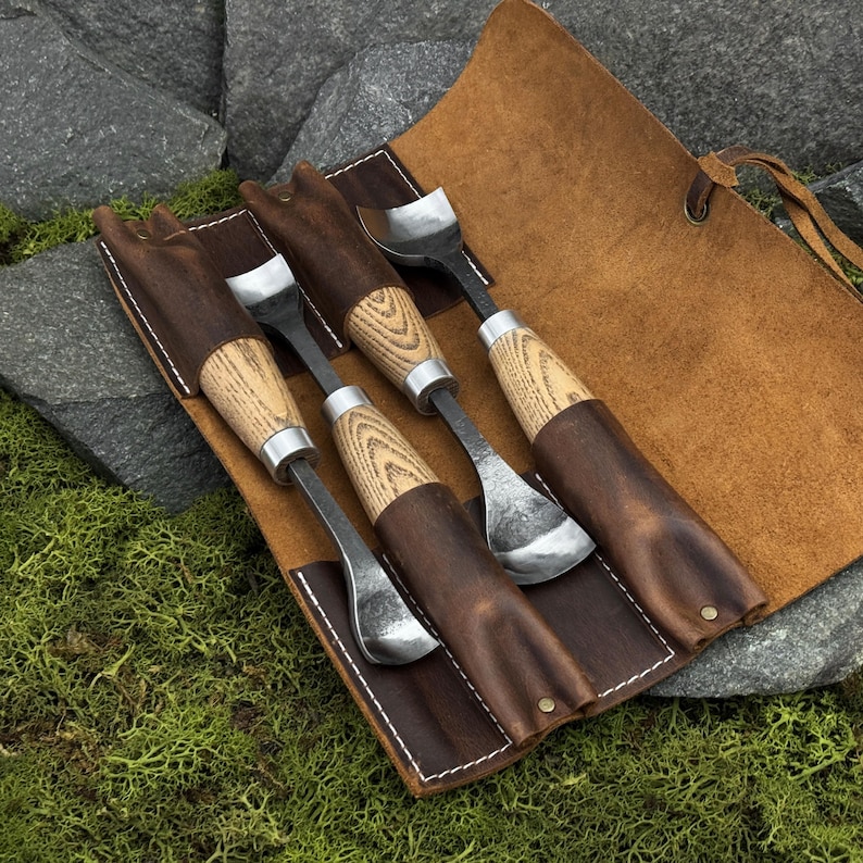 May include: A set of three metal carving tools with light-colored wooden handles, each featuring a curved blade. The tools are stored in a brown leather roll-up case with white stitching, designed with individual slots for each tool.