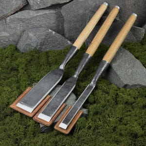 Puede incluir: Tres cinceles con mangos de madera y hojas de metal, cada uno descansando sobre una funda de cuero marrón. Las hojas son de color plateado y los mangos son de color marrón claro. Los cinceles están dispuestos en diagonal sobre un fondo de musgo verde y rocas grises.
