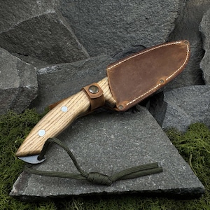 May include: A handcrafted knife with a light wood handle and a brown leather sheath. The knife features a small blade and is secured with a leather strap. A green cord is attached to the handle.