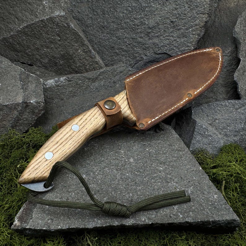 May include: A handcrafted knife with a light wood handle and a brown leather sheath. The knife features a small blade and is secured with a leather strap. A green cord is attached to the handle.