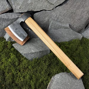 Peut inclure: Une hache forgée à la main avec un manche en bois et un protège-lame en cuir. La tête de la hache est gris foncé et le manche est brun clair. Le protège-lame en cuir est de couleur beige. La hache repose sur un lit de mousse verte et de pierres grises.