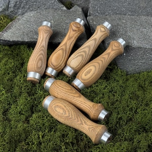Può includere: Sei manici per scalpello in legno con ghiere in metallo argentato. I manici sono in legno marrone chiaro con un evidente motivo a venature. Sono disposti su un letto di muschio verde, con rocce grigie sullo sfondo. I manici sono per utensili da falegname.
