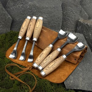 May include: A set of six wood carving gouges with curved metal blades and light-colored wooden handles with visible grain, arranged on a tan leather tool roll with a tie closure. The tools feature silver-toned metal ferrules at the handle base.