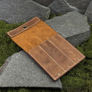 May include: Open, brown leather pen case with white stitching and a snap closure. The case has three interior slots and a flap to secure pens. The leather has a distressed look, and the case is set against a background of rocks and moss.