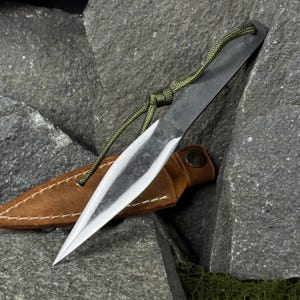Peut inclure: Un couteau de lancer forgé à la main avec une lame argentée tranchante et un manche texturé foncé. Il est doté d'un cordon vert pour la prise en main et est accompagné d'un étui en cuir marron. Le couteau repose sur des roches grises.