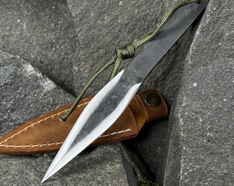 May include: A hand-forged throwing knife with a sharp, silver blade and a dark, textured handle. It features a green cord for grip and is paired with a brown leather sheath. The knife rests against a backdrop of gray rocks.