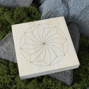 May include: A square, light-colored wooden block with a geometric design etched into its surface. The design features a central point with radiating lines and geometric shapes, creating a symmetrical pattern. The block is resting on a bed of green moss and gray stones.