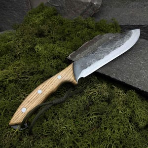 May include: A handcrafted knife with a light wood handle and a dark, textured blade. The handle is secured with three silver rivets. A green cord is attached to the base of the handle. The knife rests on a bed of green moss and a dark gray stone.