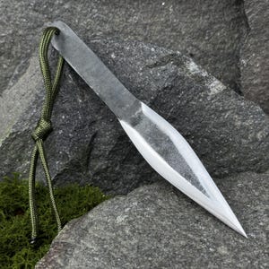 May include: A hand-forged, steel throwing knife with a sharpened blade and a lanyard made of green cord. The knife is resting on a gray rock surface, showcasing its sharp point and overall design.