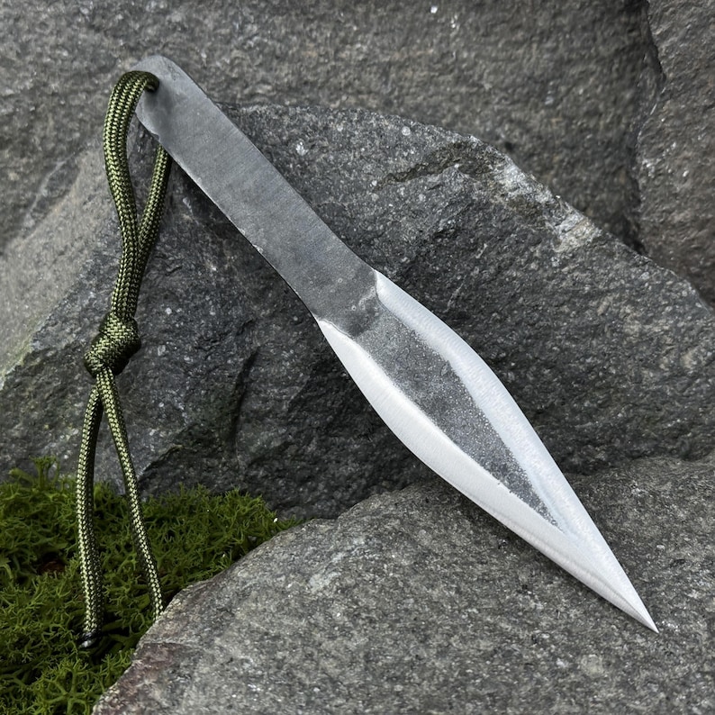 May include: A hand-forged, steel throwing knife with a sharpened blade and a lanyard made of green cord. The knife is resting on a gray rock surface, showcasing its sharp point and overall design.
