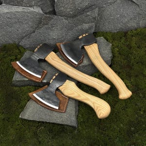 Può includere: Quattro piccole asce con manici in legno e lame in metallo scuro. Ogni ascia ha un fodero in pelle marrone con cuciture bianche. Le asce sono disposte su muschio verde e rocce grigie.