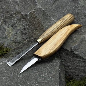 Puede incluir: Dos herramientas para tallar madera con hojas de metal. Una herramienta tiene una hoja recta y un mango cilíndrico de madera con una férula de latón. La otra herramienta tiene una hoja curva y un mango curvo de madera. Las herramientas están sobre una superficie de piedra gris.