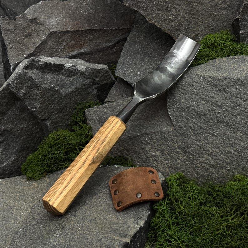 May include: A metal gouge tool featuring a curved, polished steel blade and a light brown, straight-grained wooden handle. Beside the tool lies a small, rectangular brown leather guard secured with six metal rivets, showcasing a functional design.