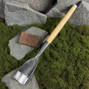 Puede incluir: Un cincel metálico grande con mango de madera y funda de cuero. El cincel tiene una hoja ancha y plana y un mango largo y cilíndrico. La funda es marrón y tiene varios ojales. El cincel y la funda descansan sobre musgo verde y piedras grises.