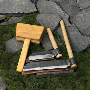 Peut inclure: Un maillet en bois avec une tête rectangulaire et un manche cylindrique, accompagné de deux outils à main dotés de lames en métal sombre, de manches en bois et de fourreaux en cuir marron, disposés sur une surface texturée.