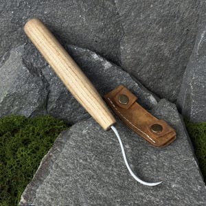 May include: A wood-handled hoof pick with a curved metal end, next to a small, brown leather sheath with two brass-colored snaps. The pick and sheath are displayed on a gray stone surface.