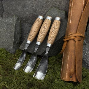 May include: Three wood carving gouges with curved, forged steel blades and light-colored wooden handles featuring visible grain patterns. Each tool has a silver metal ferrule connecting the blade to the handle, accompanied by a brown leather roll.