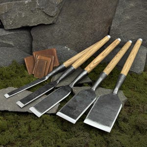 Puede incluir: Un juego de cinco cinceles de madera con mangos de madera clara y hojas de metal oscuro. Los cinceles están dispuestos sobre un lecho de musgo verde, con una pila de pequeños artículos de cuero marrón en el fondo.