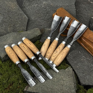 May include: A set of eight wood carving chisels with wooden handles and metal blades. The chisels are arranged on a stone surface with a leather tool roll in the background. The handles have a light wood grain pattern and are capped with silver metal.