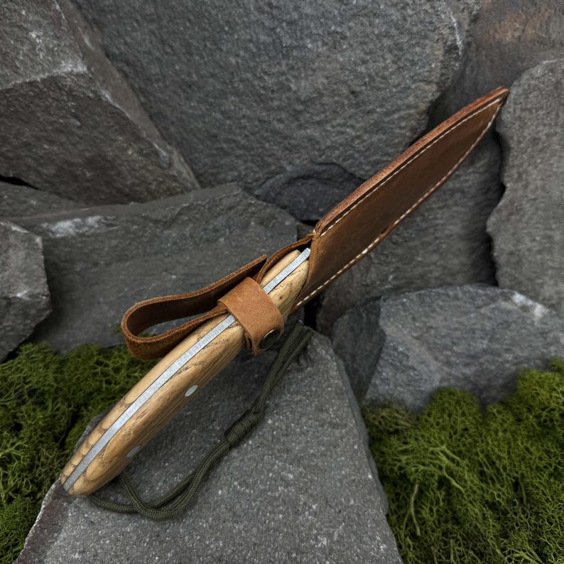 May include: A handcrafted knife with a light wood handle and a silver blade, encased in a brown leather sheath. The sheath has a loop for belt wear and is secured with a leather strap. A green cord is attached to the sheath.