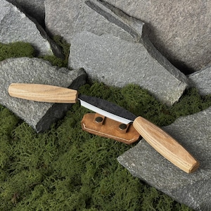 May include: A handcrafted knife with a light wood handle and a dark blade, resting on a brown leather sheath. The knife is displayed against a backdrop of gray stones and green moss, suggesting an outdoor or camping theme.