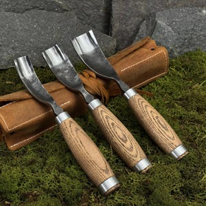 May include: Three wood carving gouges with curved, polished steel blades and light brown wooden handles. Each tool features a silver metal ferrule at the base of the handle. The gouges are arranged in a row, resting on a brown leather tool roll.