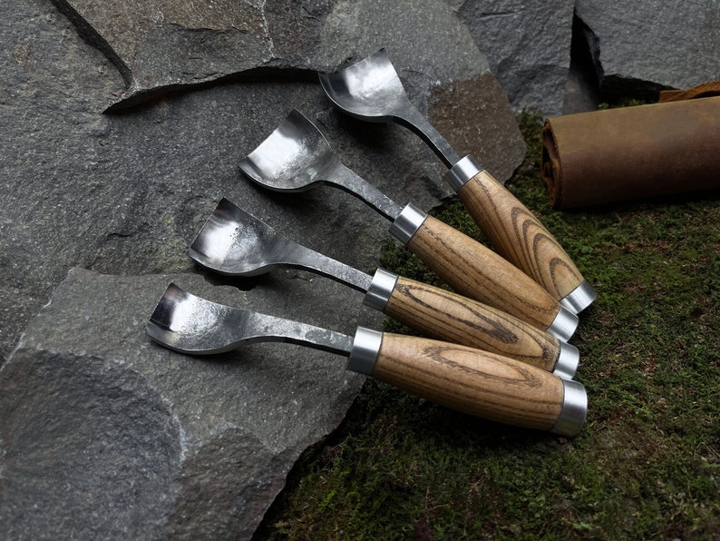 May include: Four wood carving tools with wooden handles and metal blades. The tools are arranged in a row on a grey rock surface with green moss in the background.