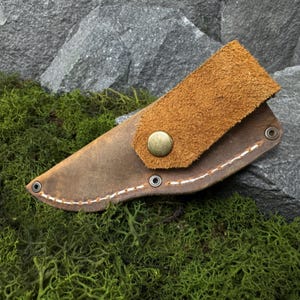 May include: A brown leather knife sheath with a suede flap secured by a brass button. The sheath is stitched with white thread and has metal eyelets. The background features a rock and green moss.