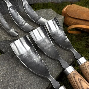 May include: A set of hand-forged metal gouges with curved, textured steel blades and wooden handles. The tools feature a dark, hammered finish on the concave metal surfaces and are arranged alongside a brown leather tool roll for storage.