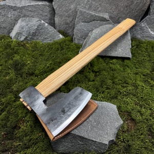 Peut inclure: Une hache artisanale avec un manche en bois clair et une tête en métal foncé. La tête de la hache repose sur un étui en cuir marron. La hache est exposée sur un lit de mousse verte et de pierres grises.