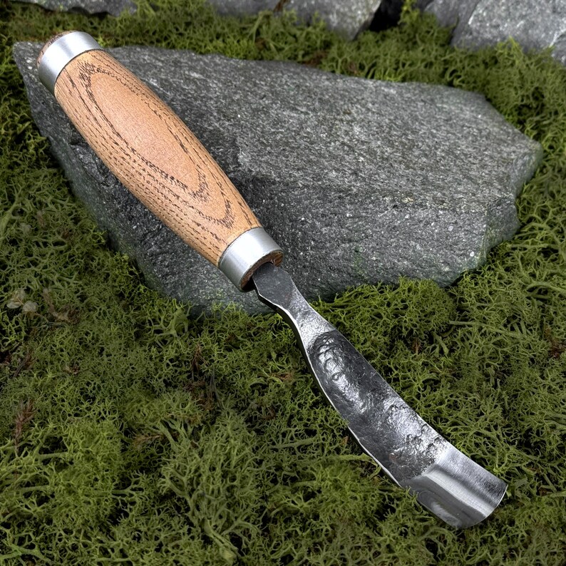 May include: A hand-forged hook knife featuring a curved, textured steel blade with a dark finish and a smooth, light-brown wooden handle secured by two polished silver-colored metal ferrules at the top and bottom of the grip.