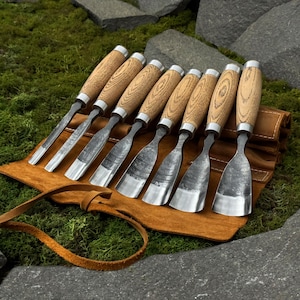 May include: A set of eight wood carving chisels with wooden handles and silver-colored blades. The chisels are arranged on a brown leather tool roll with a leather strap. The background features green moss and gray stones.