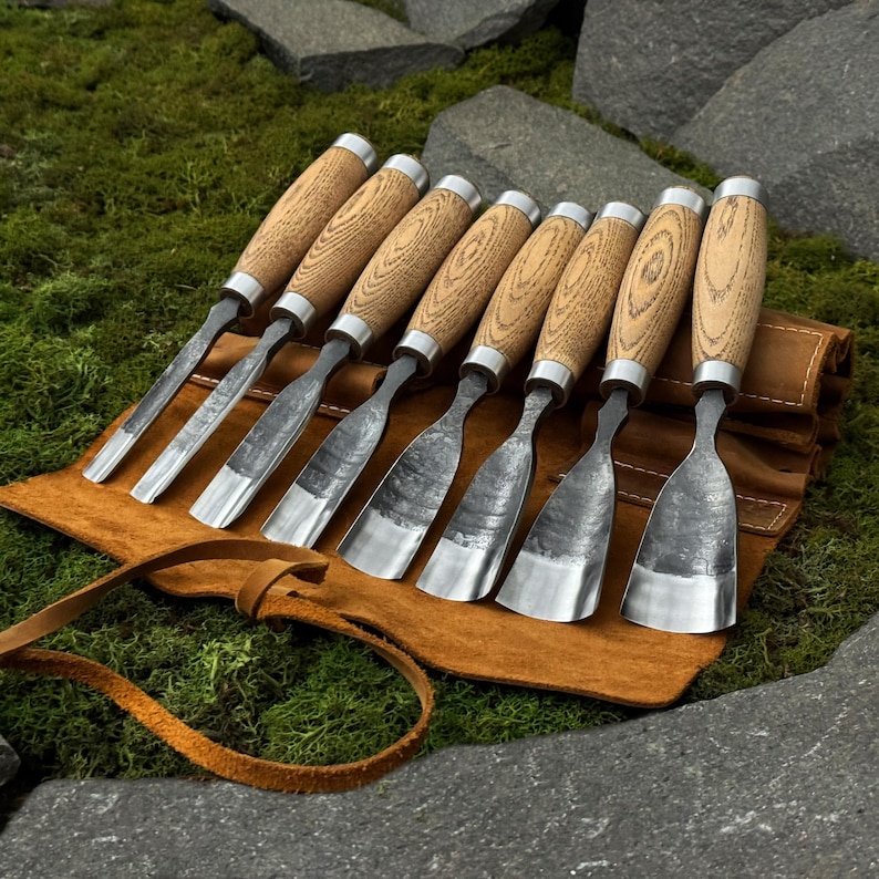 May include: A set of eight wood carving chisels with wooden handles and silver-colored blades. The chisels are arranged on a brown leather tool roll with a leather strap. The background features green moss and gray stones.